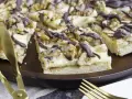Ein "friss dich dumm kuchen" mit cremiger Füllung, Walnüssen und Schokoladendrizzle auf einem Holztablett. Goldene Gabel und Messer liegen daneben.