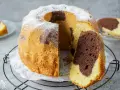 Ein saftiger Marmorkuchen Gugelhupf, mit Puderzucker bestäubt, wartet auf seinen Anschnitt. Ein Stück ist bereits abgetrennt.