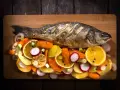 Eine ganze Forelle im Backofen, serviert mit bunten Gemüsescheiben wie Karotten, Radieschen und Orangen auf einem Holzbrett.