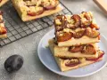 Saftiger zwetschgenkuchen vegan mit Streuseln auf einem Teller gestapelt. Daneben frische Zwetschgen und ein Kuchenblech.