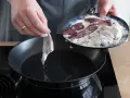 Ein Stück Leber wird in eine heiße Pfanne mit Öl gelegt, um sie zu braten. Auf einem Teller liegen weitere Leberstücke, die in Mehl gewendet wurden.