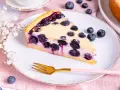Ein Stück Heidelbeerkuchen Landfrauenart mit saftigen Blaubeeren und cremigem Guss auf einem Teller, daneben eine goldene Gabel und frische Beeren.