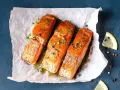 Drei saftige **Lachs im Ofen**-Portionen, garniert mit Kräutern und Pfeffer, auf Backpapier. Daneben Zitronenspalten.