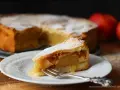 Ein Stück gedeckter Apfelkuchen mit Puderzucker bestäubt, auf einem Teller mit einer Gabel. Im Hintergrund der ganze Kuchen und Äpfel.
