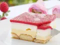 Himbeer-Sahne-Schnitten auf weißer Platte, mit frischen Himbeeren und rosa Rosen im Hintergrund. Ein Stück wird mit einer Gabel herausgenommen.