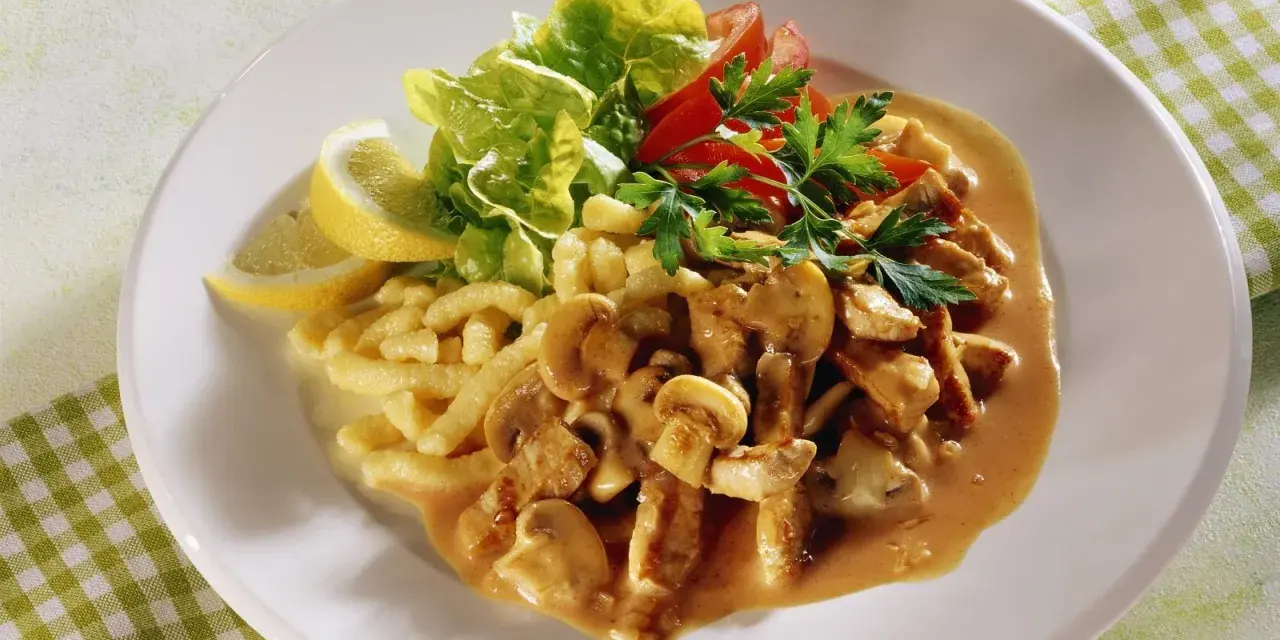 Ein Teller mit geschnetzeltem Fleisch mit Pilzen in cremiger Sauce, dazu Spätzle, Salat und Zitronenspalten.