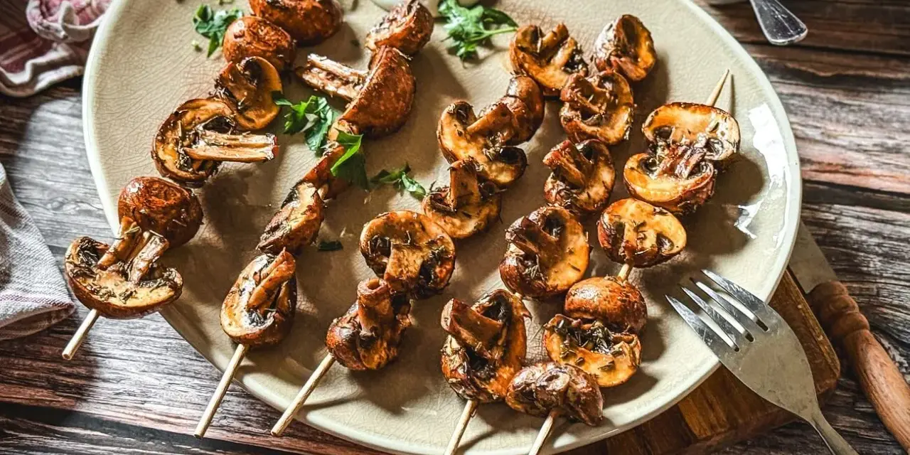 Gegrillte Champignons auf Spießen, mit Kräutern verfeinert und auf einem Teller mit Dip serviert. Ein Genuss für jeden Pilzliebhaber!