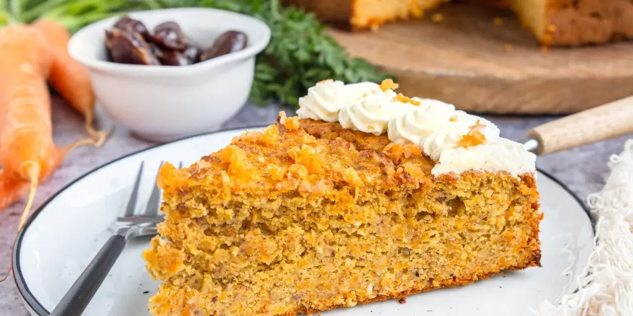 Ein saftiges Stück karottenkuchen ohne zucker, verziert mit cremiger Glasur und Orangenschale. Daneben liegen frische Karotten und Datteln.