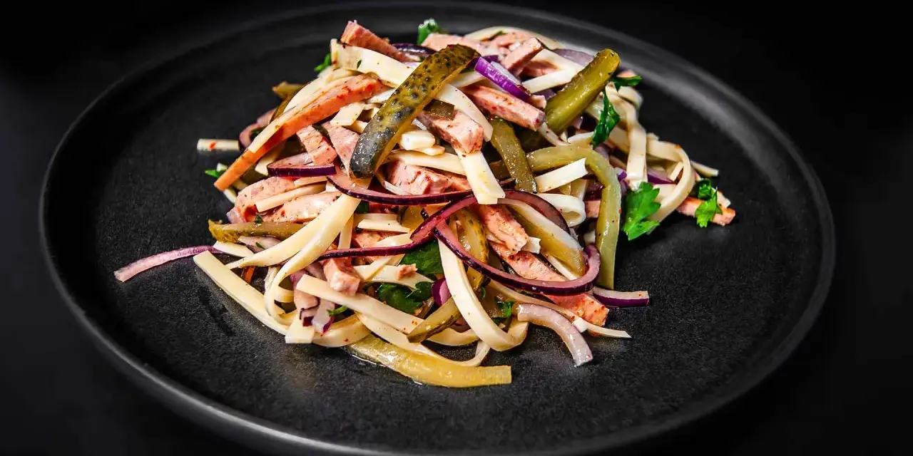 Ein appetitlicher Schweizer Wurstsalat mit Wurststreifen, Käse, roten Zwiebeln und Gewürzgurken auf einem dunklen Teller.