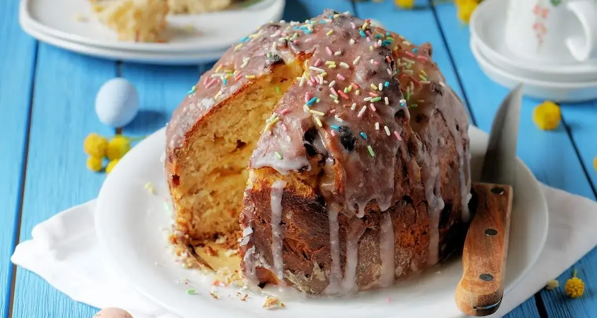 Ein Anschnitt eines Paska-Rezepts mit Zuckerguss und bunten Streuseln. Daneben liegen bunte Ostereier und gelbe Blümchen.