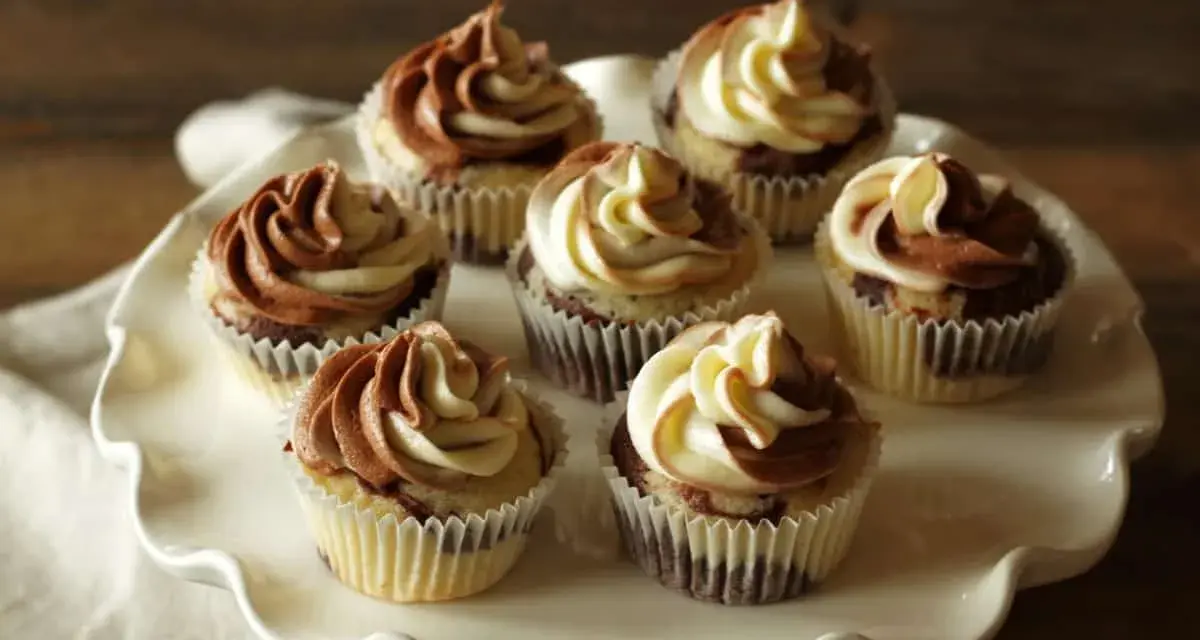 Sieben Marmormuffins mit zweifarbiger Schokoladen-Vanille-Glasur auf einer weißen Tortenplatte. Ein köstliches Marmormuffins Rezept.