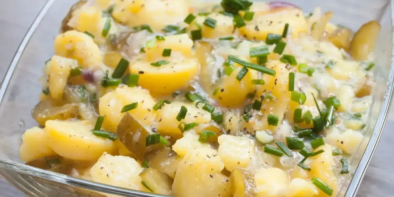 Ein köstlicher Kartoffelsalat ohne Mayo, mit Gurkenstücken und frischem Schnittlauch garniert, in einer Glasschüssel.