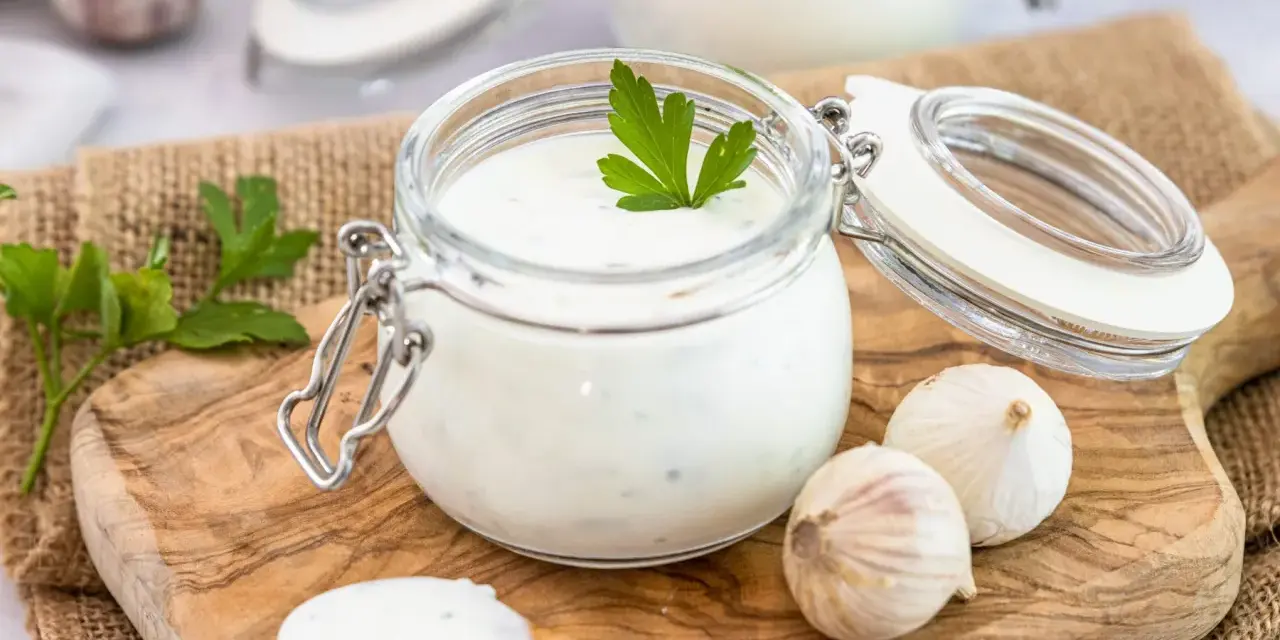Ein Glas mit cremigem Knoblauch Dip, garniert mit Petersilie, daneben Knoblauchzehen und ein Löffel mit Dip.