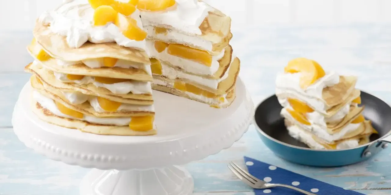 Eine köstliche Pfannkuchen Torte mit Pfirsichen und Sahne, auf einem weißen Tortenständer und ein Stück in einer kleinen Pfanne.