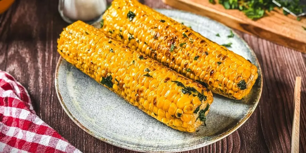 Zwei goldgelbe maiskolben, perfekt gegrillt und mit Kräutern bestreut, liegen auf einem Teller. Ein Genuss für alle, die maiskolben grillen lieben.