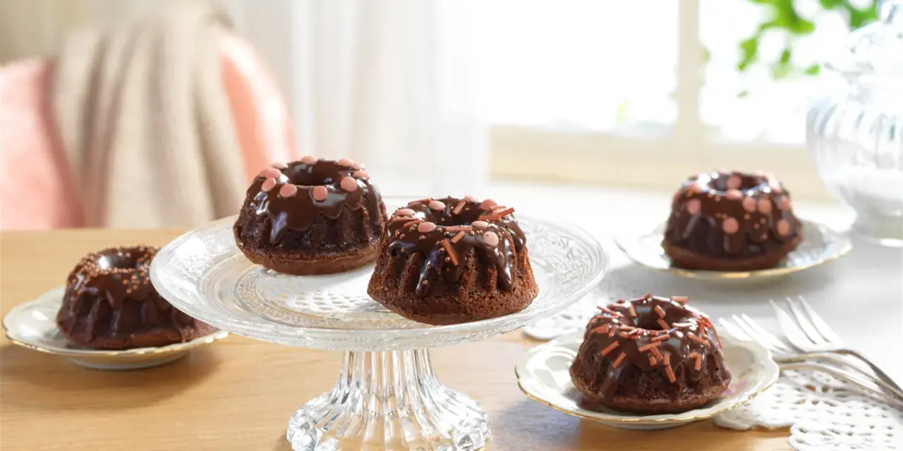 Schokoladen-Mini-Gugelhupf dekorieren mit Schokoglasur und bunten Streuseln auf einem Tortenständer.