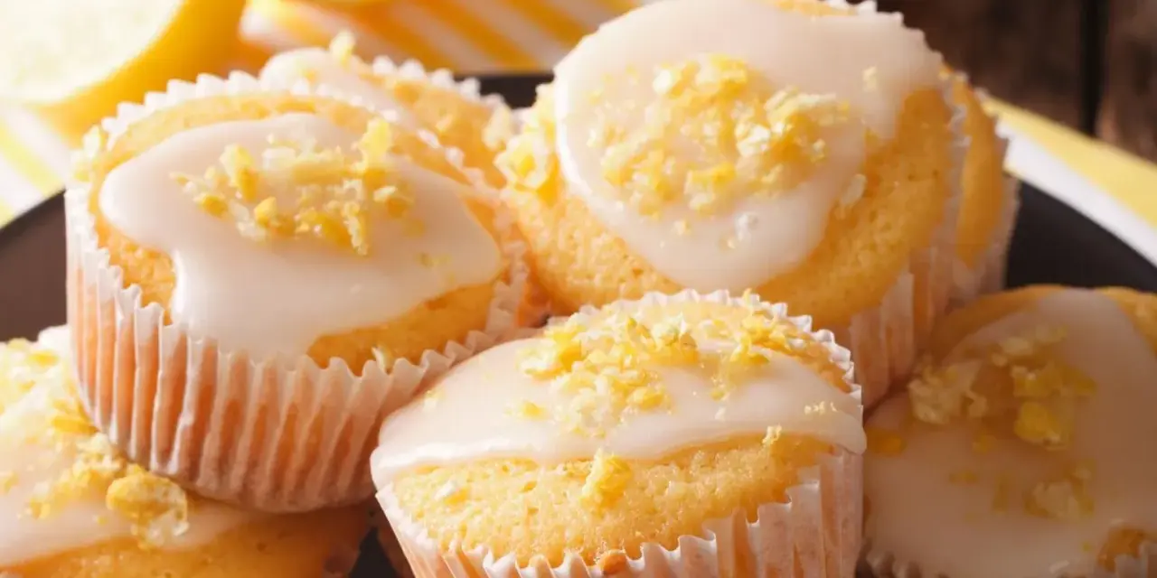Leckere zitronenmuffins rezept: goldgelbe Muffins mit weißer Glasur und Zitronenschale bestreut, daneben frische Zitronenscheiben.