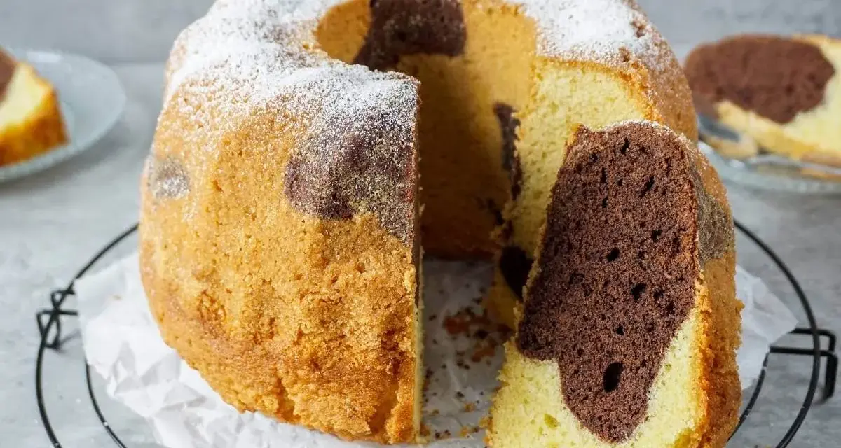Ein saftiger Marmorkuchen Gugelhupf, mit Puderzucker bestäubt, wartet auf seinen Anschnitt. Ein Stück ist bereits abgetrennt.