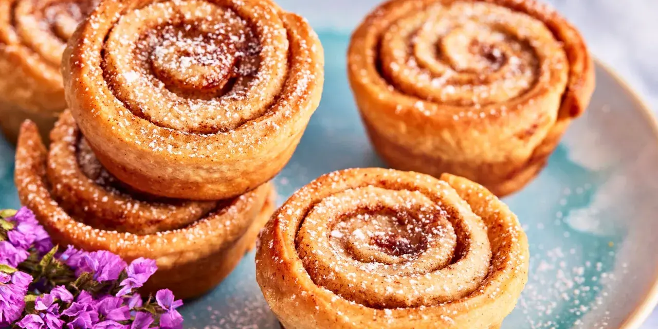 Saftige Zimtschnecken Muffins, bestäubt mit Puderzucker, liegen auf einem blauen Teller. Lila Blumen schmücken die süße Leckerei.