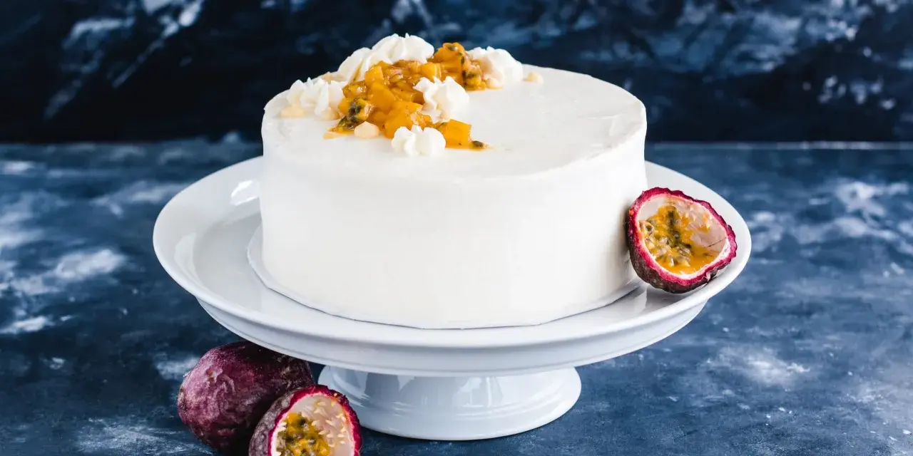 Eine weiße Maracuja Torte mit frischer Maracuja-Frucht und Sahnetupfern, umgeben von ganzen und halbierten Maracujas.