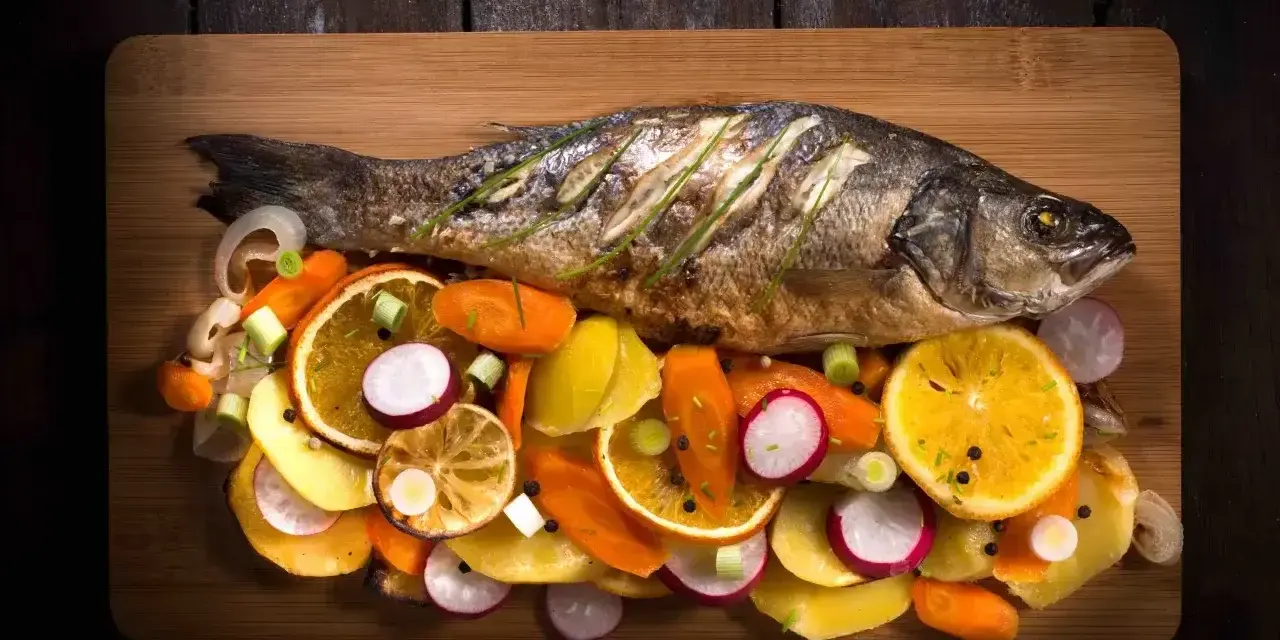 Eine ganze Forelle im Backofen, serviert mit bunten Gemüsescheiben wie Karotten, Radieschen und Orangen auf einem Holzbrett.