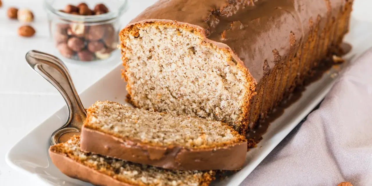 Saftiger Mohn Nuss Kuchen mit Schokoladenglasur, daneben ein Glas mit Haselnüssen und zwei angeschnittene Scheiben.