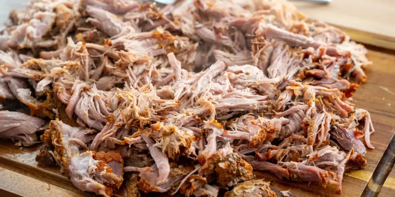Ein Haufen zartes, **gezupftes Fleisch** liegt auf einem Holzbrett, bereit für leckere Sandwiches oder Tacos. Eine Gabel liegt daneben.
