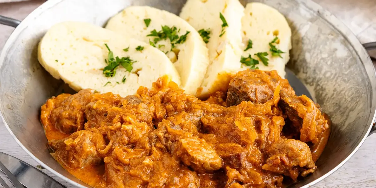 Herzhaftes Szegediner Gulasch mit Klößen, garniert mit Petersilie, serviert in einer Metallschüssel.