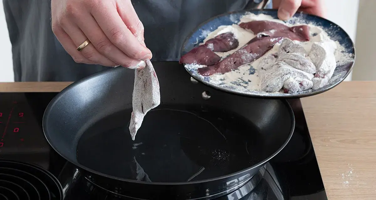 Ein Stück Leber wird in eine heiße Pfanne mit Öl gelegt, um sie zu braten. Auf einem Teller liegen weitere Leberstücke, die in Mehl gewendet wurden.