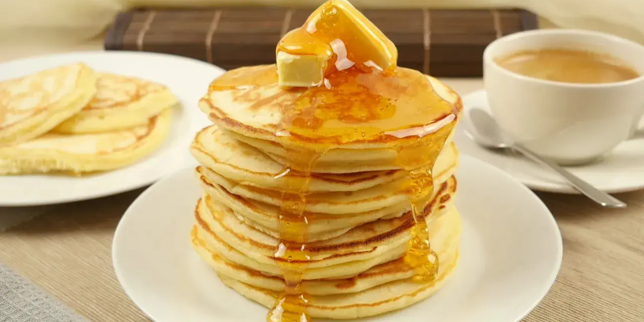 Ein Stapel goldbrauner Pancakes, übergossen mit Honig und Butter, daneben eine Tasse Kaffee. Perfekt für ein leckeres Pancakes Rezept.