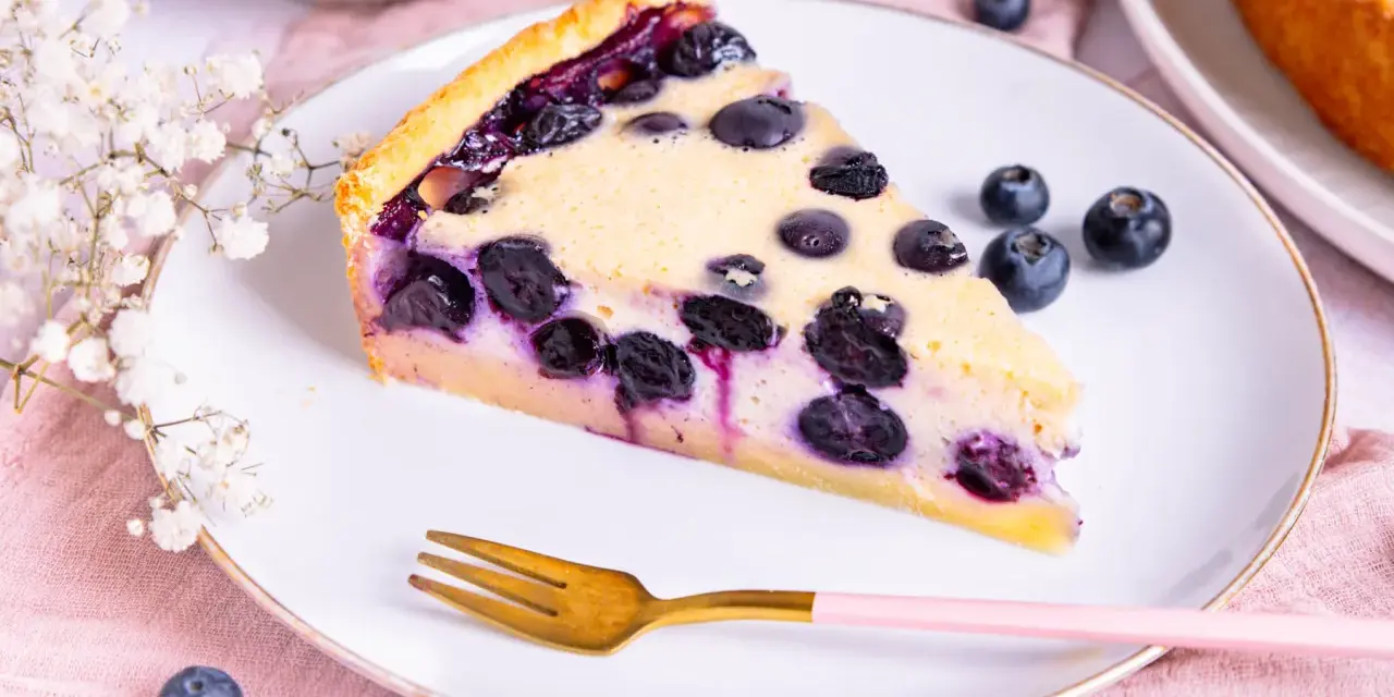 Ein Stück Heidelbeerkuchen Landfrauenart mit saftigen Blaubeeren und cremigem Guss auf einem Teller, daneben eine goldene Gabel und frische Beeren.