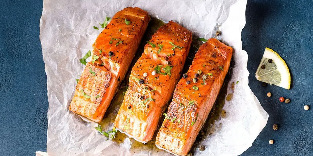 Drei saftige **Lachs im Ofen**-Portionen, garniert mit Kräutern und Pfeffer, auf Backpapier. Daneben Zitronenspalten.