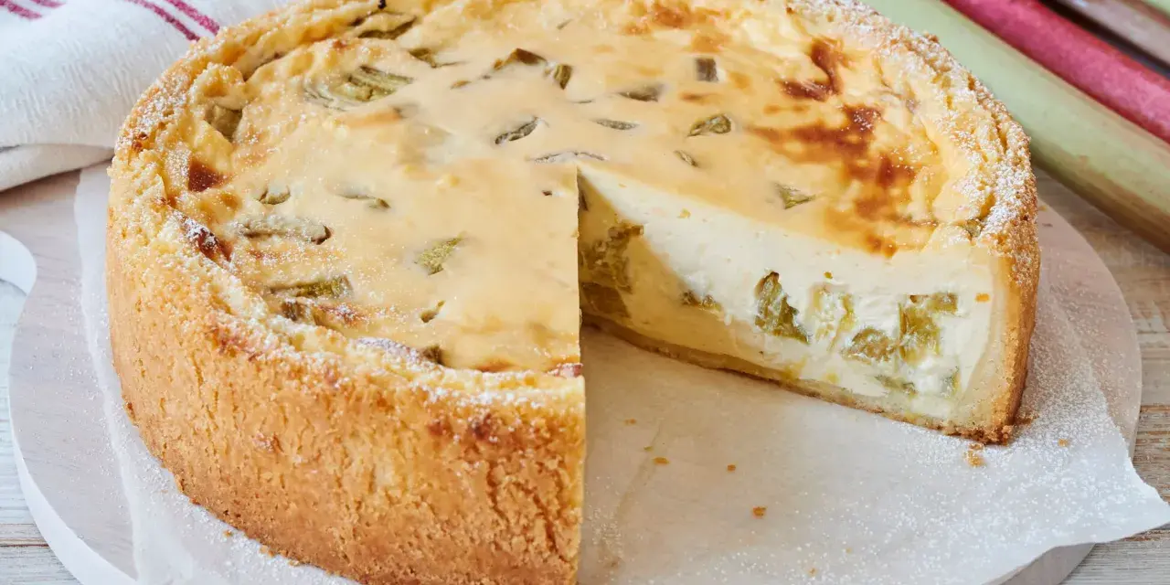 Ein saftiger Rhabarberkuchen, schnell gebacken und mit Puderzucker bestäubt. Daneben liegen frische Rhabarberstangen.