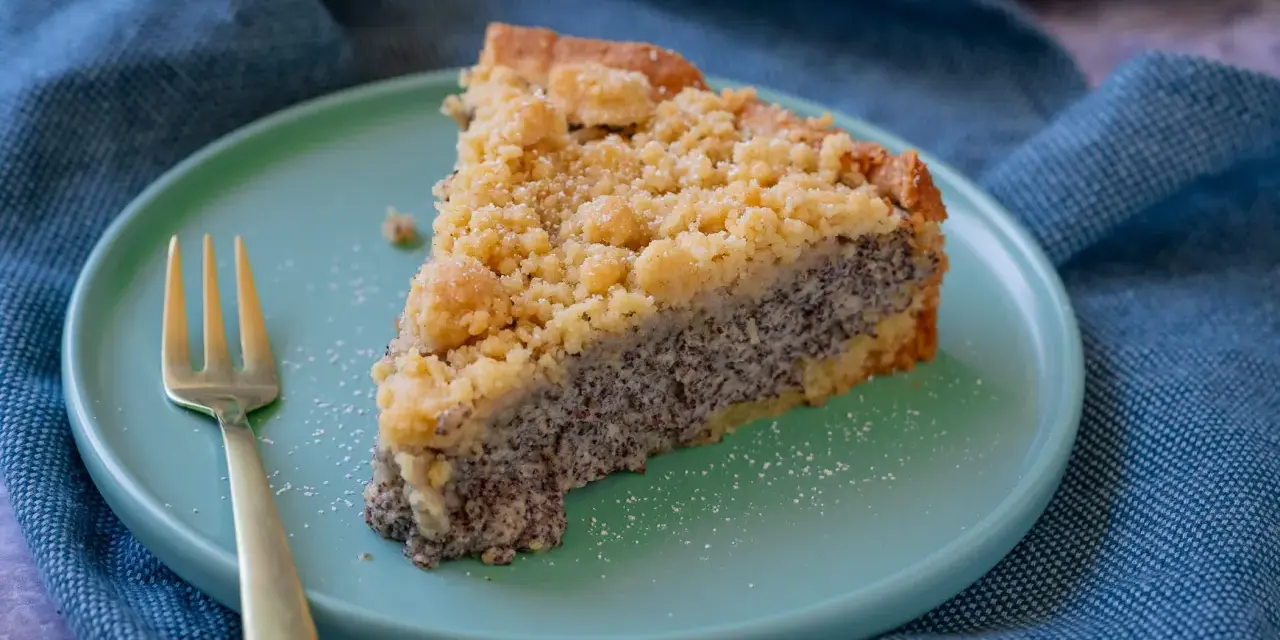 Ein saftiges Stück Mohnkuchen mit Streuseln auf einem mintgrünen Teller, daneben eine goldene Gabel. Ein köstliches Mohnkuchen Rezept.