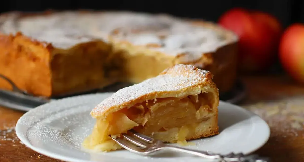 Ein Stück gedeckter Apfelkuchen mit Puderzucker bestäubt, auf einem Teller mit einer Gabel. Im Hintergrund der ganze Kuchen und Äpfel.