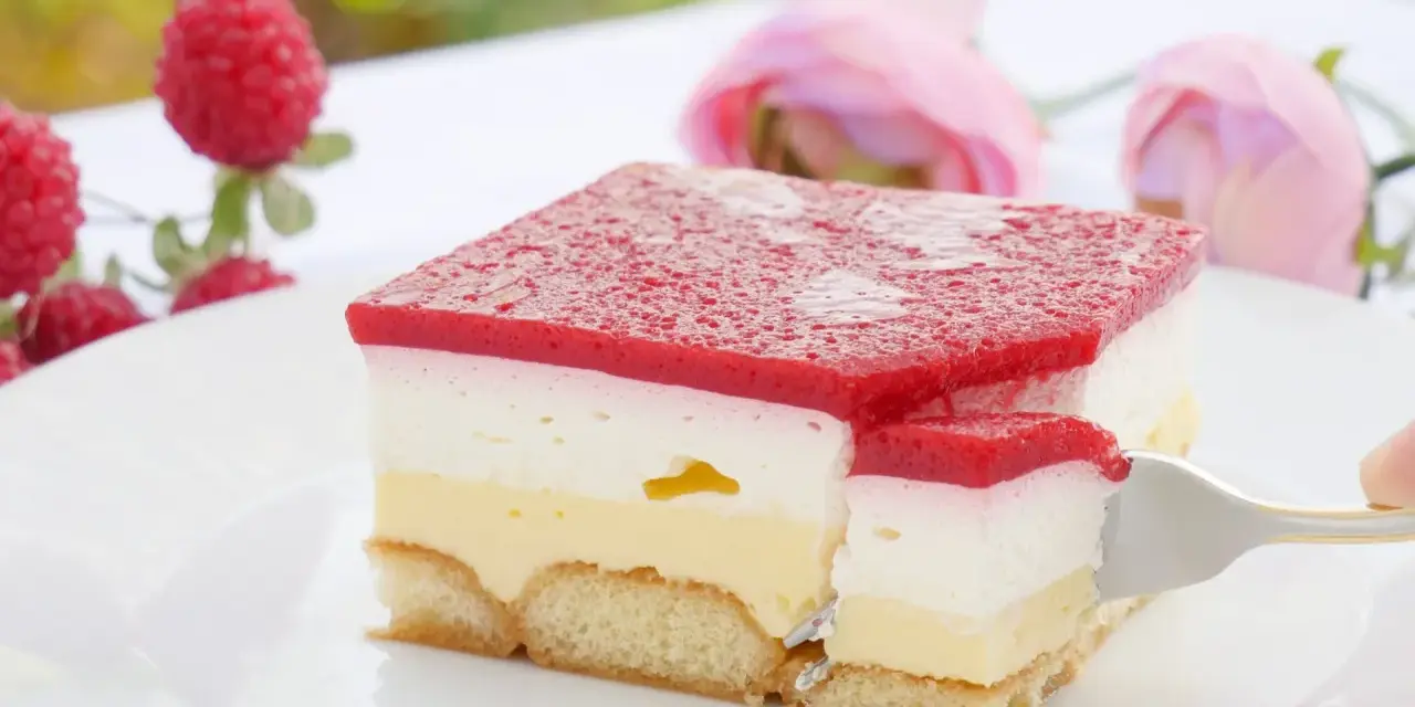 Himbeer-Sahne-Schnitten auf weißer Platte, mit frischen Himbeeren und rosa Rosen im Hintergrund. Ein Stück wird mit einer Gabel herausgenommen.
