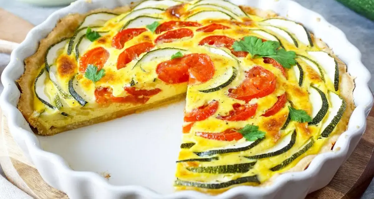 Eine köstliche Zucchini-Quiche mit Tomaten und frischer Petersilie, in einer weißen Quicheform, ein Stück fehlt.
