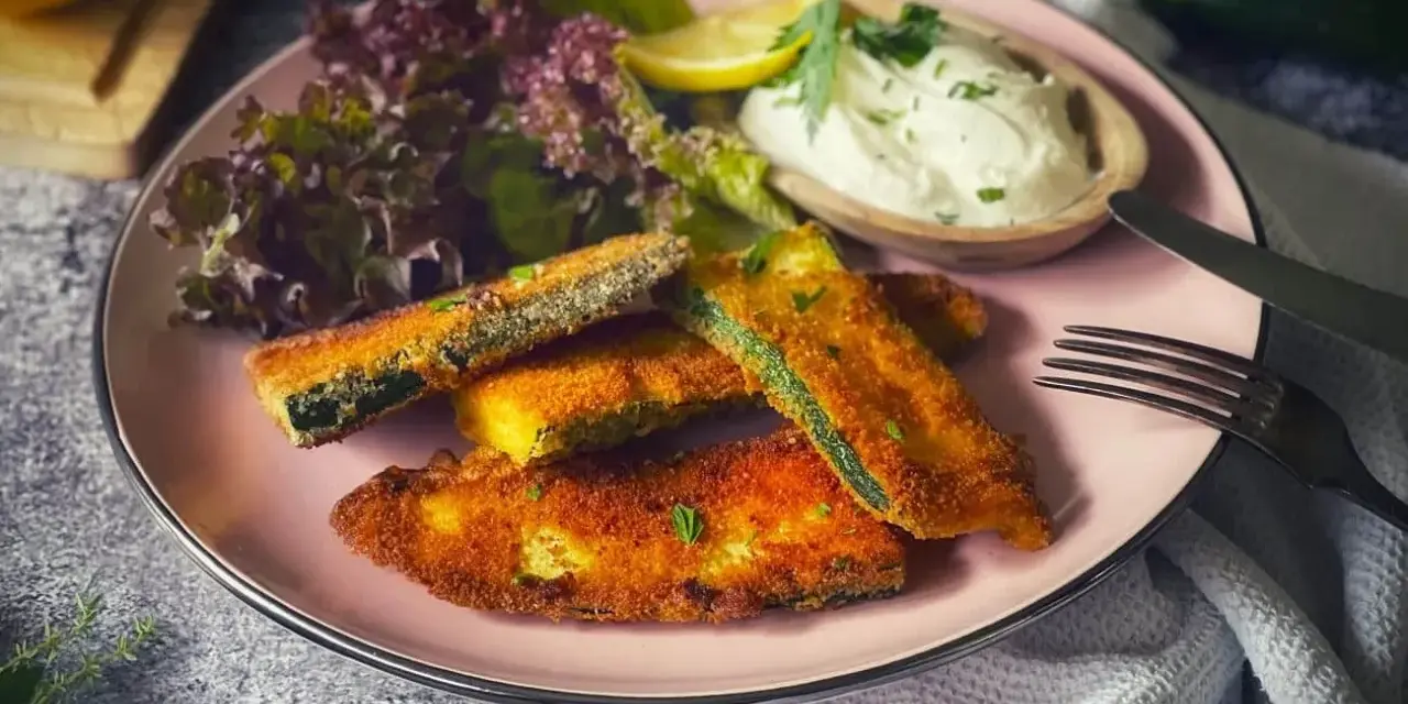 Knuspriges Zucchini-Schnitzel auf rosa Teller, serviert mit Dip, Salat und Zitrone. Ein Genuss für jeden Tag!