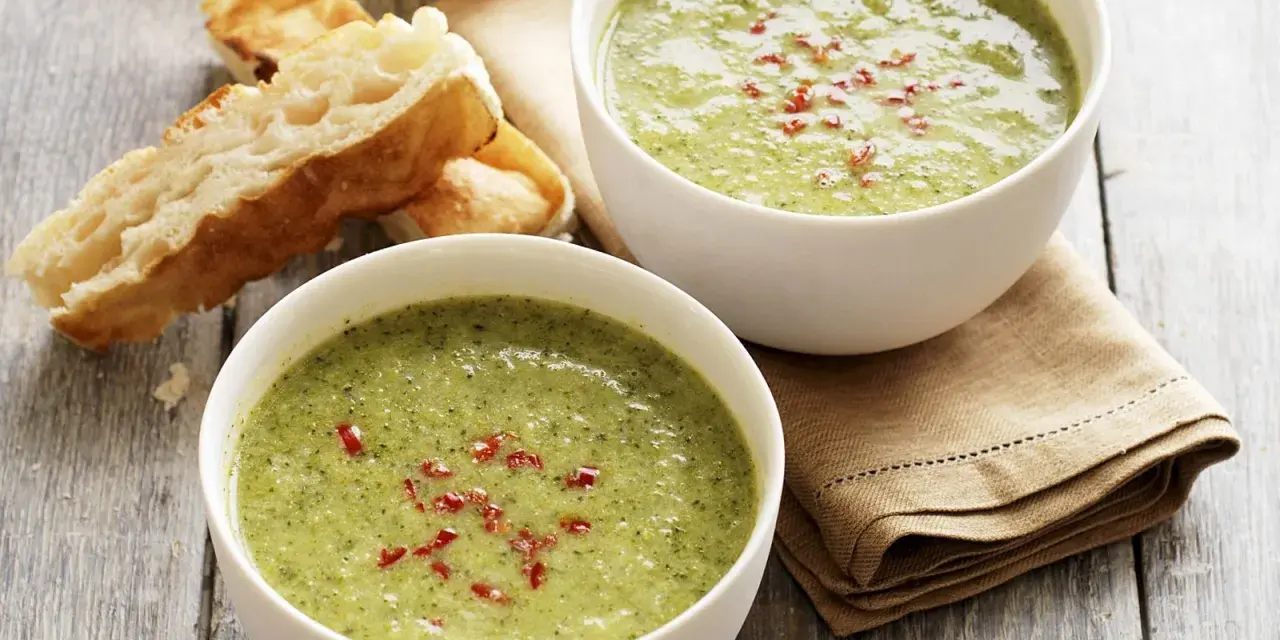 Zwei Schalen cremiger Brokkolisuppe, garniert mit roten Chiliflocken, stehen neben knusprigem Baguette auf einem Holztisch.
