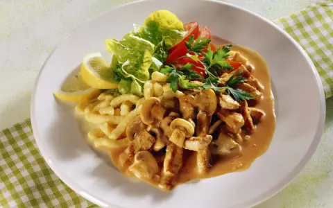 Ein Teller mit geschnetzeltem Fleisch mit Pilzen in cremiger Sauce, dazu Spätzle, Salat und Zitronenspalten.