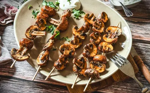 Gegrillte Champignons auf Spießen, mit Kräutern verfeinert und auf einem Teller mit Dip serviert. Ein Genuss für jeden Pilzliebhaber!
