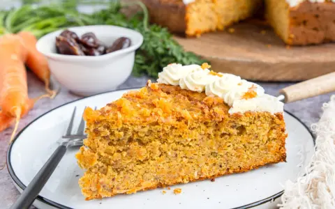 Ein saftiges Stück karottenkuchen ohne zucker, verziert mit cremiger Glasur und Orangenschale. Daneben liegen frische Karotten und Datteln.