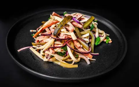 Ein appetitlicher Schweizer Wurstsalat mit Wurststreifen, Käse, roten Zwiebeln und Gewürzgurken auf einem dunklen Teller.