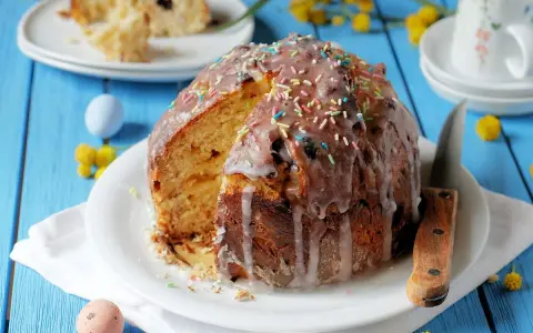 Ein Anschnitt eines Paska-Rezepts mit Zuckerguss und bunten Streuseln. Daneben liegen bunte Ostereier und gelbe Blümchen.