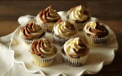 Sieben Marmormuffins mit zweifarbiger Schokoladen-Vanille-Glasur auf einer weißen Tortenplatte. Ein köstliches Marmormuffins Rezept.