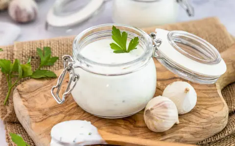 Ein Glas mit cremigem Knoblauch Dip, garniert mit Petersilie, daneben Knoblauchzehen und ein Löffel mit Dip.