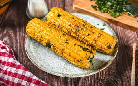 Zwei goldgelbe maiskolben, perfekt gegrillt und mit Kräutern bestreut, liegen auf einem Teller. Ein Genuss für alle, die maiskolben grillen lieben.