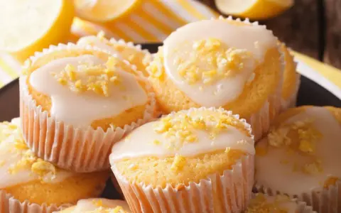 Leckere zitronenmuffins rezept: goldgelbe Muffins mit weißer Glasur und Zitronenschale bestreut, daneben frische Zitronenscheiben.