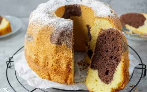 Ein saftiger Marmorkuchen Gugelhupf, mit Puderzucker bestäubt, wartet auf seinen Anschnitt. Ein Stück ist bereits abgetrennt.