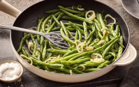 Grüne Bohnen mit Zwiebelringen in einer Pfanne, bereit zum Kochen. Daneben eine Schale mit grobem Salz.